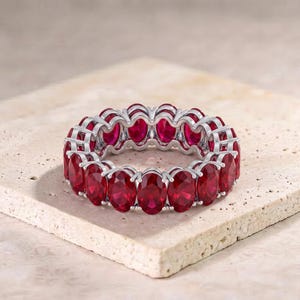 May include: An eternity band ring featuring a continuous row of oval-cut vibrant red gemstones set in a polished silver-toned metal setting. The ring displays a uniform arrangement of stones around the entire circumference of the band.