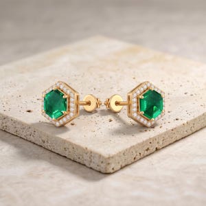May include: A pair of stud earrings crafted in yellow gold, each featuring a central hexagonal-cut green emerald surrounded by a halo of small, round, clear white diamonds. The earrings include standard post backs for secure attachment.