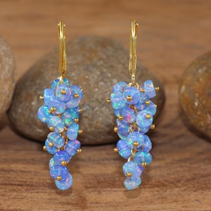 Könnte beinhalten: Ohrringe mit Anhängern aus hellblauen Opalperlen. Die Perlen haben schillernde Farbtöne und sind an goldfarbenen Klappbügelverschlüssen befestigt. Die Ohrringe werden vor einem Hintergrund aus Naturholz und Stein präsentiert.