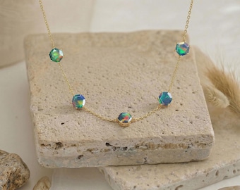 May include: A delicate gold necklace featuring five iridescent, hexagonal gemstones. The gemstones display a range of colors, including green, blue, and purple. The necklace is displayed on a textured, light-colored surface.