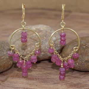 以下が含まれることがあります： ピンク色の宝石クラスターが付いたゴールドトーンのフープピアス。ピアスは円形のデザインで、小さなファセット加工されたピンク色の宝石が繊細なチェーンから吊り下げられており、レイヤード効果を生み出しています。 レバーバッククロージャーを備えています。