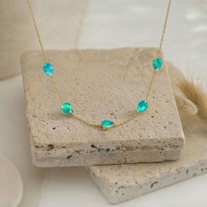 Könnte beinhalten: Eine zarte goldene Halskette mit fünf tropfenförmigen Opalen in Blau- und Grüntönen. Die Opale sind gleichmäßig auf einer feinen Goldkette angeordnet. Die Halskette wird auf einer strukturierten, hellfarbigen Oberfläche präsentiert.