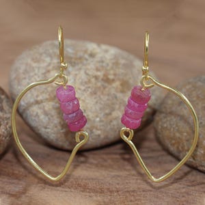Könnte beinhalten: Ein Paar goldfarbene Ohrringe mit herzförmigem Design. Jeder Ohrring hat eine Ansammlung kleiner, rosa, facettierter Perlen. Die Ohrringe werden vor einem natürlichen, neutral getönten Hintergrund präsentiert.