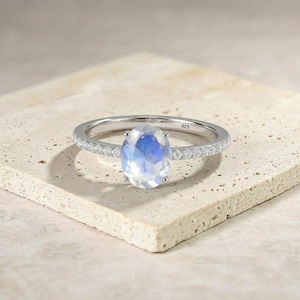 Könnte beinhalten: Ein silberner Ring mit einem großen, oval geformten, irisierenden Edelstein. Das Band ist mit kleinen, klaren Steinen besetzt. Der Ring liegt auf einer strukturierten, neutral getönten Oberfläche, die das Design und die Handwerkskunst des Schmucks hervorhebt.
