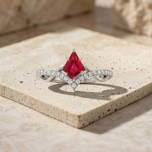 Könnte beinhalten: Silberring mit einem facettierten, roten Edelstein in Drachenform als Mittelpunkt. Der Ring hat ein Band mit kleinen, klaren Edelsteinen und einem dekorativen, wirbelnden Design. Der Ring liegt auf einer strukturierten Oberfläche in neutralen Farbtönen.