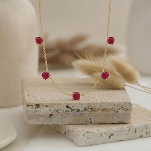 May include: A delicate gold necklace featuring six square-cut, vibrant red gemstones. The necklace is displayed on a textured, light-colored stone surface, with a neutral background. The chain is fine, and the gemstones are evenly spaced.