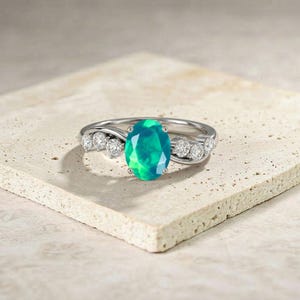 Könnte beinhalten: Ein silberner Ring mit einem oval geschliffenen, leuchtend grünen Opal. Der Ring ist mit kleinen, runden, klaren Edelsteinen besetzt. Der Ring liegt auf einer neutralfarbenen Oberfläche. Der Opal misst ca. 1,5 cm x 1 cm.