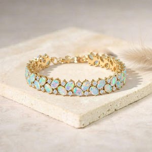 May include: A gold bracelet featuring oval-shaped opals and small, round diamonds. The opals are a mix of white, blue, and green. The bracelet is set on a textured, light-colored surface.