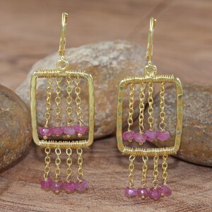 Könnte beinhalten: Goldfarbene Ohrringe mit quadratischem Rahmendesign. Jeder Ohrring hat herabhängende Ketten mit kleinen, rosa Edelsteinen. Die Ohrringe haben einen Klappverschluss. Der Hintergrund ist natürlich strukturiert.