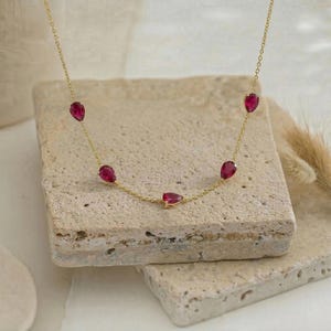May include: A delicate gold chain necklace featuring five evenly spaced, pear-shaped, deep red gemstones. The necklace is constructed with a thin, polished gold link chain that holds each teardrop-shaped stone in a secure, minimalist metal setting.