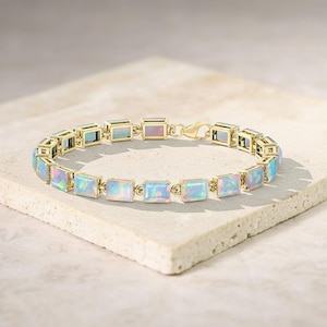 May include: A gold bracelet with rectangular opal stones. The opals display a play of colour, with blue, pink, and green hues. The bracelet is set on a neutral surface, highlighting the jewellery's design.