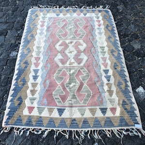 Puede incluir: Una alfombra tejida tradicional con un patrón geométrico en tonos de rosa, gris y marrón. La alfombra tiene un borde de flecos y está colocada sobre una superficie de piedra.