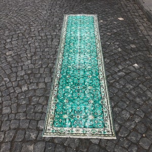 May include: A long, teal-colored rug with a white border and intricate floral patterns. The rug is laid out on a gray cobblestone surface.