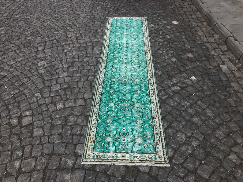 May include: A long, teal-colored rug with a white border and intricate floral patterns. The rug is laid out on a gray cobblestone surface.