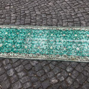 May include: A vintage teal and white patterned rug with a floral design. The rug has a fringed edge and is laid out on a gray cobblestone surface.