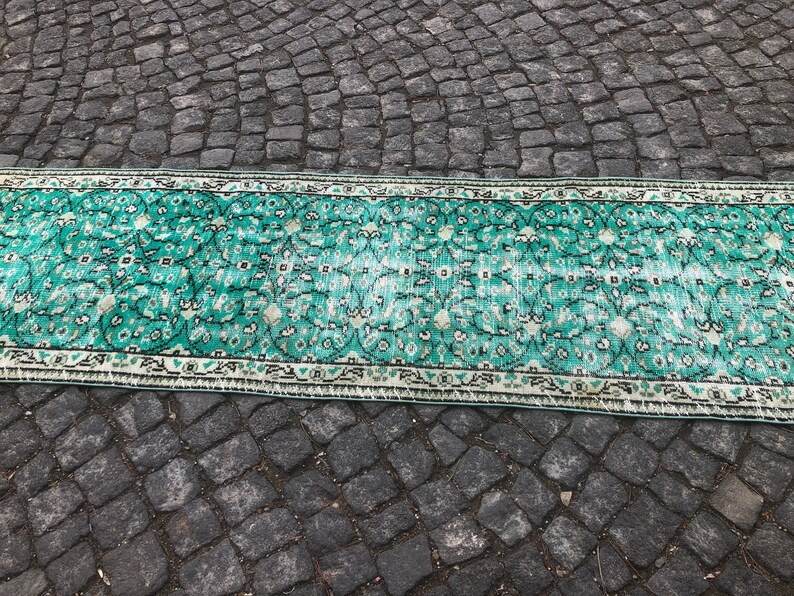May include: A vintage teal and white patterned rug with a floral design. The rug has a fringed edge and is laid out on a gray cobblestone surface.