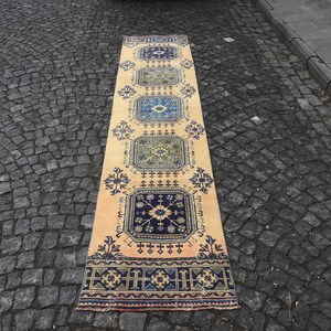 Peut inclure: Un long tapis de couleur crème avec un motif géométrique dans des tons de bleu, vert et beige. Le tapis a une bordure à franges et est posé sur une surface en pavés.