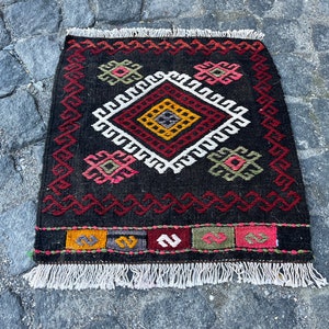 May include: A black and red rug with a geometric pattern in white, yellow, and green. The rug has a fringe border.