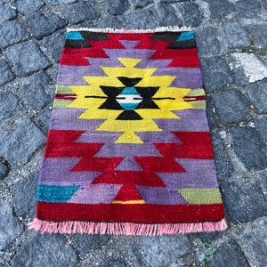 May include: A colorful geometric rug with a central diamond pattern in shades of yellow, black, and blue. The rug has a fringe border and is made of wool.