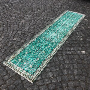 May include: A teal and white patterned runner rug with a floral design. The rug is laid out on a gray cobblestone surface.