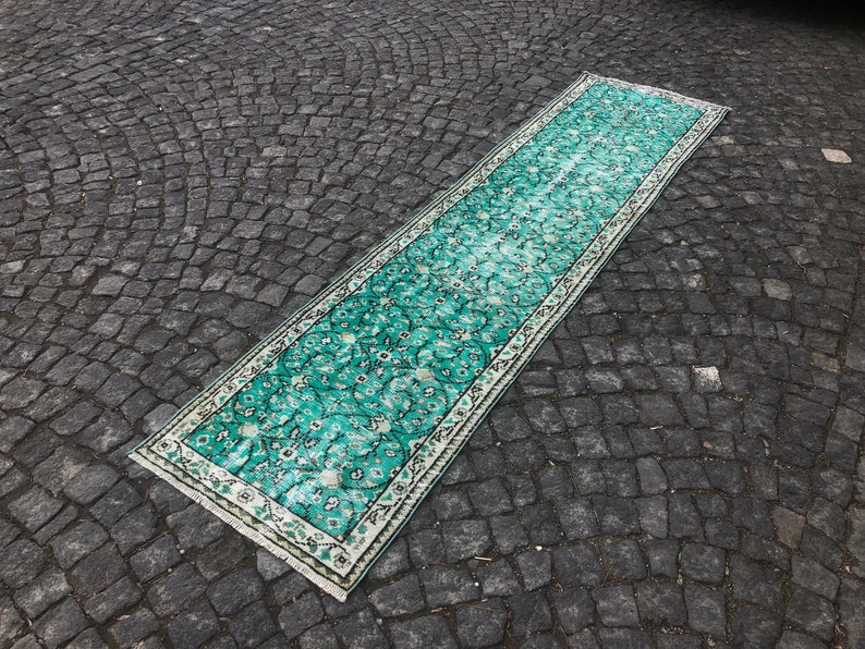 May include: A teal and white patterned runner rug with a floral design. The rug is laid out on a gray cobblestone surface.