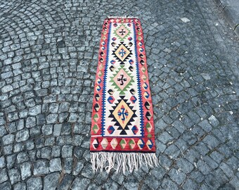 May include: A long, narrow rug with a traditional Turkish kilim design. The rug features a white background with red, blue, green, and yellow geometric patterns. The rug has fringe on both ends.