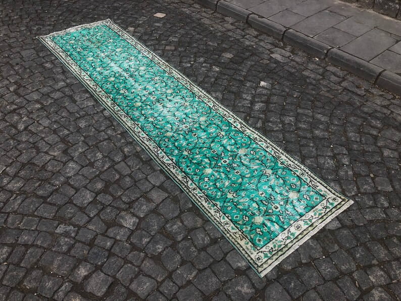 May include: A long, teal-colored rug with a white border and a floral pattern. The rug is laid out on a gray cobblestone surface.