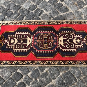 May include: A red, black, and cream colored rug with a geometric pattern. The rug has a fringe border and is made of wool.