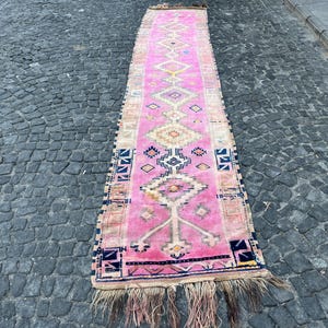 May include: A long, rectangular pink rug with a geometric pattern. The rug features a central design of diamonds and other shapes in white, yellow, and blue. Fringes are visible at the ends. The rug is likely made of wool or a similar material.