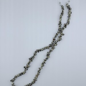 May include: A long eyeglass chain constructed from numerous small, irregular, tumbled dalmatian jasper stone beads featuring a cream base with black and dark brown spots. The chain terminates at both ends with clear silicone loops and metal coils.
