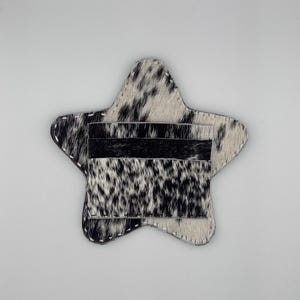 May include: A star-shaped card holder made of cowhide with a black and white spotted pattern. The card holder has three black card slots and white stitching around the edges. The overall design is a unique and stylish accessory.