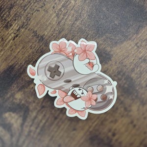 May include: A sticker featuring a gray video game controller design with pink cherry blossoms and a cartoon character. The character is white with its tongue sticking out. The controller has a cross-shaped directional pad and several buttons.
