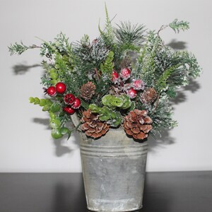 May include: A festive arrangement of artificial greenery, red berries, and pine cones in a galvanized metal bucket. The arrangement includes frosted pine branches, green leaves, and several brown pine cones. The bucket has a handle and a rustic, weathered appearance.