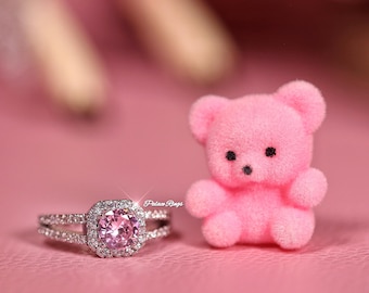 May include: A pink gemstone ring with a square halo setting and a split band sits next to a small pink teddy bear on a pink surface. The ring features a large, round pink stone surrounded by smaller clear stones. The teddy bear has black eyes and a black nose.
