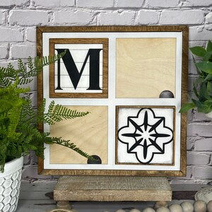 May include: A wooden sign with a white background and a brown frame. The sign is divided into four squares. The top left square has a black letter "M" on a white background. The top right square is blank. The bottom left square is blank. The bottom right square has a black and white geometric design. The sign is sitting on a wooden stand.