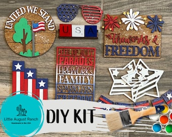 Könnte beinhalten: Ein DIY-Set zum Basteln von patriotischen Dekorationen. Das Set enthält Holzausschnitte mit roter, weißer und blauer Farbe, einen Pinsel und eine Reihe von Farbtöpfen. Die Ausschnitte zeigen Sätze wie "United We Stand", "Fireworks & Freedom" und "4th of July Parades".