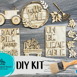 May include: DIY craft kit with wooden cutouts for fall decorations. The kit includes a tractor, a rake, a pile of leaves, a wagon, a wreath with the words "hello fall", and a sign that says "unbe leaf able". The kit also includes paint and a paintbrush.