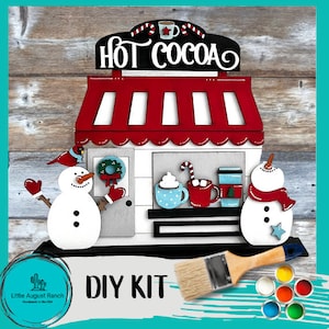 May include: DIY craft kit featuring a wooden cutout of a hot cocoa stand with snowmen, a paintbrush, and paint pots. The stand has a red awning with the words "Hot Cocoa" on the front. The kit includes everything needed to create a festive winter scene.