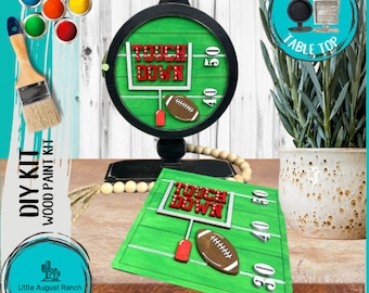 May include: DIY wood paint kit featuring a football field with a goal post and the words "Touch Down". The kit includes a round wooden sign and a rectangular wooden sign. The signs are painted green and white with red and brown accents. The kit also includes a set of paint colors.