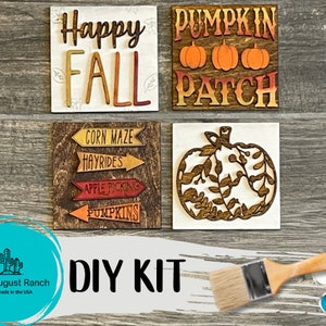 May include: DIY craft kit featuring four wooden signs with fall-themed designs. The signs include "Happy Fall", "Pumpkin Patch", a pumpkin with leaves, and a sign with arrows that read "Corn Maze", "Hayrides", "Apple Picking", and "Pumpkins". The kit includes paint and a paintbrush.
