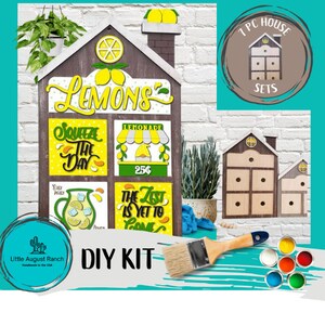 May include: DIY kit for a wooden lemonade stand house with a lemon theme. The kit includes four pre-cut wooden panels with painted designs, a paintbrush, and a set of acrylic paints. The panels feature the words "Lemons", "Squeeze the Day", "Lemonade 25¢", and "The Zest is Yet to Come".