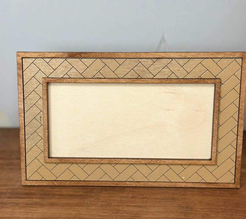 K&ouml;nnte beinhalten: Ein Holzrahmen mit hellbrauner Oberfl&auml;che und einem goldfarbenen geometrischen Muster. Der Rahmen hat eine rechteckige &Ouml;ffnung f&uuml;r ein Foto oder ein Kunstwerk.