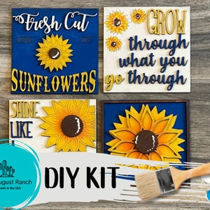 May include: DIY craft kit featuring four wooden signs with sunflower designs and inspirational quotes. The signs are painted blue and yellow and include the phrases "Fresh Cut Sunflowers", "Grow through what you go through", "Shine Like", and a large sunflower. The kit includes paint and a paintbrush.