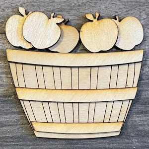 May include: Wooden craft cutout of a basket filled with apples. The basket is brown and has a wood grain texture. The apples are white and have a smooth surface.