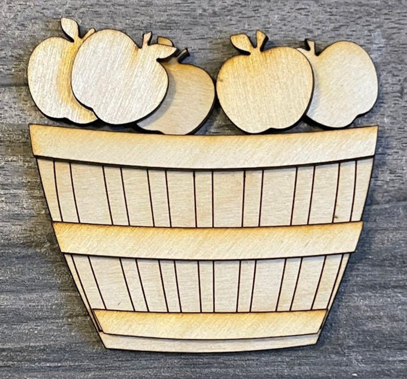 May include: Wooden craft cutout of a basket filled with apples. The basket is brown and has a wood grain texture. The apples are white and have a smooth surface.