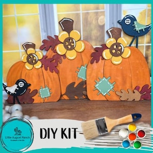 May include: DIY craft kit featuring three wooden pumpkins with fall leaves, a bird, and a paintbrush. The pumpkins are painted in orange, yellow, and brown. The kit includes a set of acrylic paints in various colors.