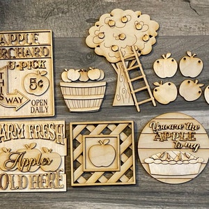 May include: A set of wooden craft cutouts for an apple orchard theme. The set includes a sign that says "Apple Orchard U-Pick 5&cent; This Way Open Daily", a sign that says "Farm Fresh Apples Sold Here", a tree with apples and a ladder, a basket of apples, a square frame with an apple inside, and a round sign that says "You are the Apple to my" with a pie.