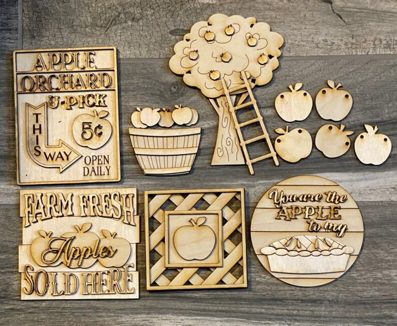 May include: A set of wooden craft cutouts for an apple orchard theme. The set includes a sign that says "Apple Orchard U-Pick 5&cent; This Way Open Daily", a sign that says "Farm Fresh Apples Sold Here", a tree with apples and a ladder, a basket of apples, a square frame with an apple inside, and a round sign that says "You are the Apple to my" with a pie.