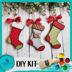 May include: DIY Christmas stocking ornament kit with three unfinished wooden stockings, paint, and a paintbrush. The stockings are decorated with red, green, and white paint. One stocking has the name "Ethan" written on it.