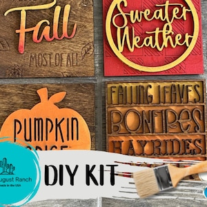 May include: DIY kit for four wooden signs with fall-themed text and images. The signs feature the text "Fall Most of All", "Sweater Weather", "Falling Leaves Bonfires Hayrides", and "Pumpkin Spice". The kit includes paint and a paintbrush.